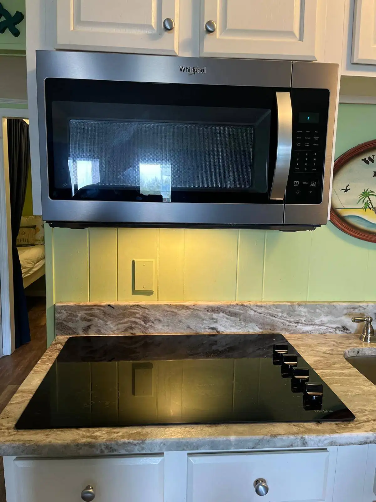 Stainless steel Whirlpool over-the-range microwave above a black glass electric cooktop in a kitchen with white cabinets and a marble-look countertop